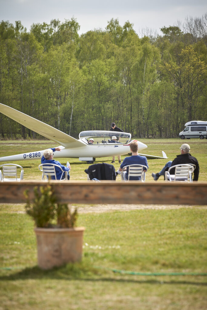 Drei Personen sitzen vor einem Segelflugzeug, in dem zwei weitere Personen sitzen