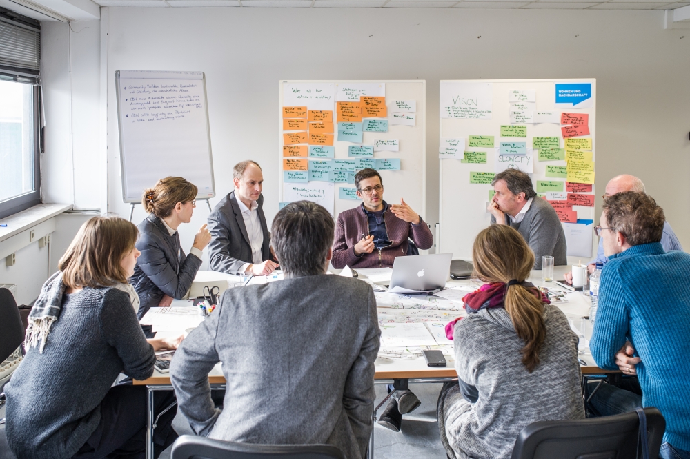 Gruppenbild mit Personen an einem Tisch, dahinter Stellwände mit Ideennotizen