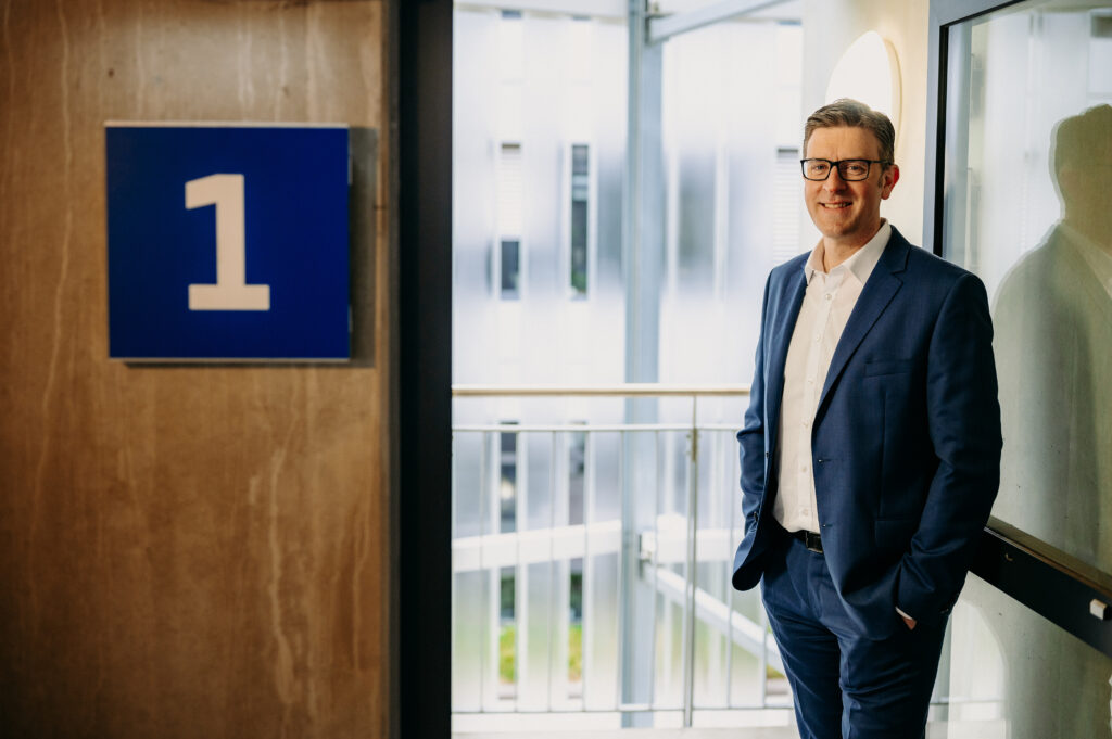 Podcast-Gast Christopher Conrads steht am Fenster, neben ihm eine große Eins an der Wand