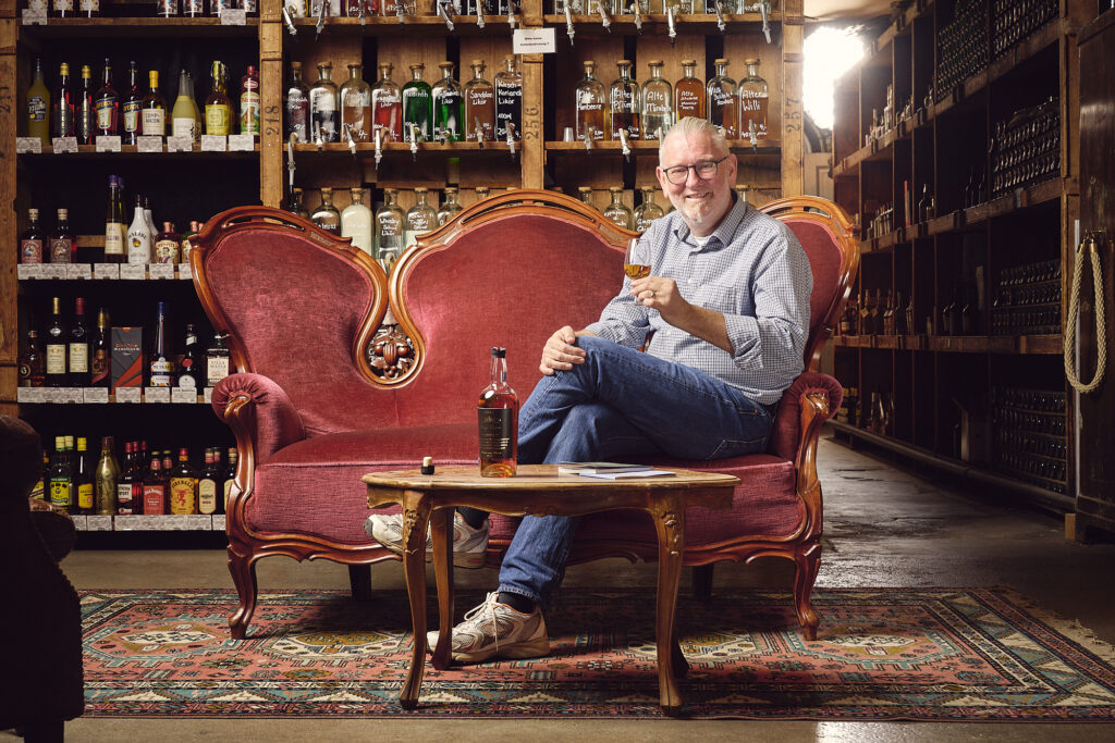 Heinrich von Have sitzt auf einem roten, alt wirkenden Sofa, in der Hand hält er ein Nosingglas, hinter ihm ein Holzregal mit Flaschen
